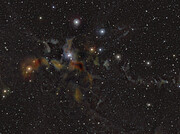 This image shows stars and clouds of gas and dust distributed over a dark background. A prominent cloud of gas and dust can be seen in the central part of the image. It features amorphous clouds in a red and brown hue. In the upper half of the image are a dozen stars in blue, red and yellow colours that shine more prominently than the other stars distributed uniformly in the image.