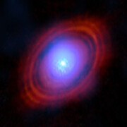 On a dark background, a bright bluish-white object at the centre of the image is surrounded by reddish rings in a disc shape. The object and rings are hazy and slightly blend.