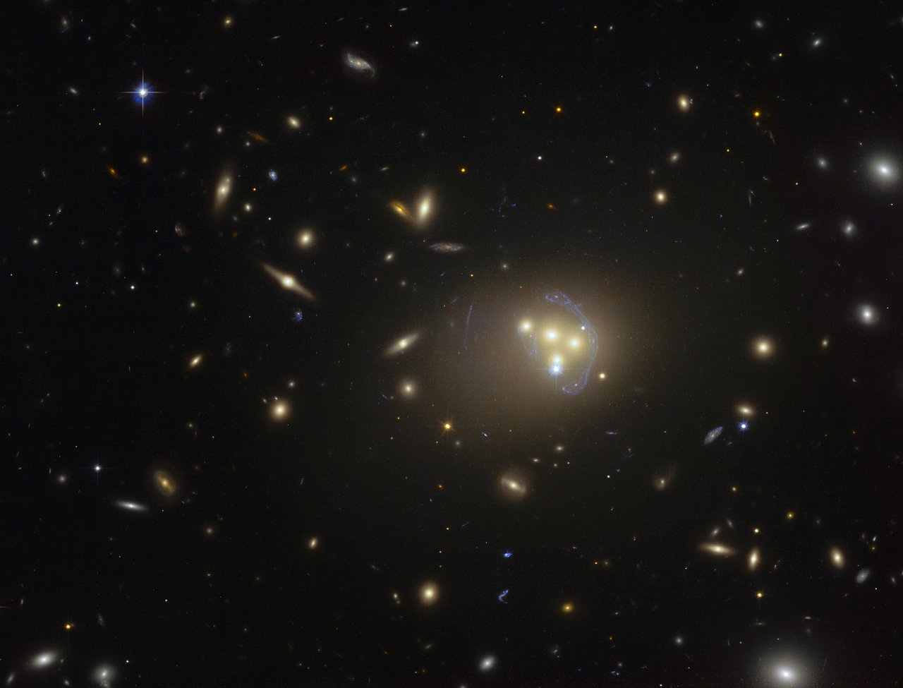 Esta imagen del telescopio espacial Hubble de NASA/ESA muestra el rico cúmulo de galaxias Abell 3827. Las extrañas estructuras azules alrededor de las galaxias centrales son una galaxia distante, situada detrás del cúmulo, vista con el efecto de lente gravitacional. Las observaciones de las cuatro galaxias centrales fusionándose han proporcionado indicios de que la materia oscura que rodea a una de las galaxias no se mueve con la propia galaxia, lo cual posiblemente implica que están teniendo lugar interacciones de naturaleza desconocida en la materia oscura con otra materia oscura.