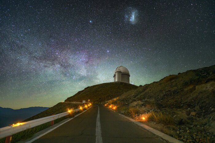 At the centre of this image two domes sit atop a mountain. The larger dome contains the 3.6-metre telescope of the La Silla Observatory. In the foreground, an access road leads up to the telescope. The road is lined with dim, warm lights. Above the mountain, the night sky is filled with a myriad of stars. To the upper left, the Milky Way glows in the sky in shades of white and purple. The Large Magellanic Cloud is a small blue and white blur at the very top of the image. At the bottom of the sky there is a faint green haze due to airglow.