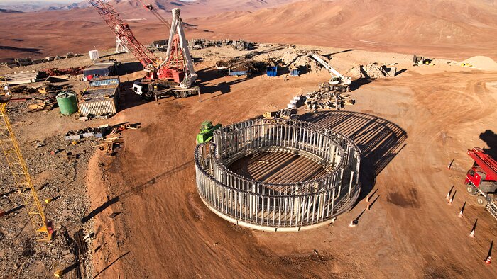 On flattened orange-brown ground sits a circular structure made up of vertical grey rods. Angled towards the back of the image, there is an array of construction equipment. Further off in the distance, the ground gives way to rolling hills.