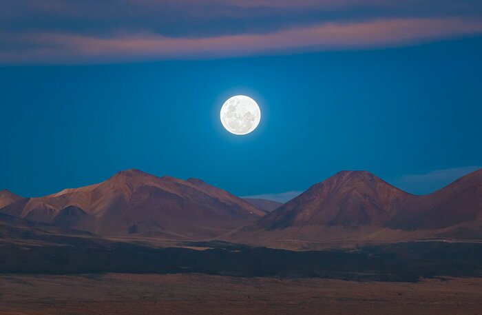 This image looks out from the Chajnantor plateau to the full Moon, glowing in white with grey patches. The Moon sits just above, and framed between, mountain peaks jutting up from the plateau, with the entire landscape in shades of rusty red and brown. The deep, yet bright, blue sky provides a contrasting background in the top half of the image.