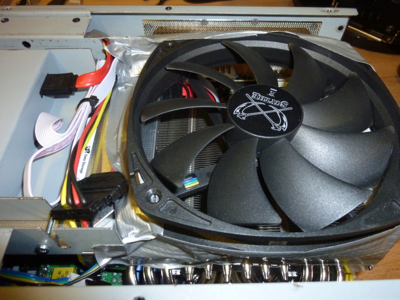 Top view w/o case lid showing rear part of card reader and the air duct with direct air flow from the fan to the GPU heatsink. The back shows the drive bay with a hole so a screw driver can get to the screw holding the card readers metal cover. To the right of that hole a slit in the drive bay has been cut for a SATA + 5V power cable. Another visible detail is the notch cut into the end of the bolt(s) that come up through the fan, which makes it easier to adjust the fan height