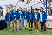 Seven people in blue ESO jackets stand outside under a white tent, smiling at the camera.