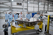 A very shiny hexagonal mirror in the foreground of the image reflects the faces of the people standing around it inside a large, white hall. The people are wearing hairnets and white or blue lab coats; some are wearing face masks and gloves too. The mirror rests on top of a yellow metallic structure, with different electronics coming out of it.
