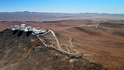This image shows a drone shot of ESO’s Paranal Observatory from high in the air. On the left-hand side of the image is a collection of large white buildings atop a mountain. A road leads down the mountain, towards other buildings in the distant desert landscape. Between the two boundaries of a large U-bend in the road, some small containers can be seen.
