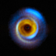 A circular hazy and colourful object sits on a black background. Towards its outside edges, it has spiral arms of blue and orange. Moving inwards, it has a brighter orange ring and a dark red inner circle. The very centre is a black blob.