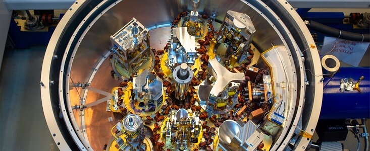 The cryostat of an ALMA antenna is at the centre of the image and occupies almost all of it. The device is an open cylindrical case made of grey metal, photographed from above. It contains a total of 10 receivers –– golden and grey metallic devices topped with funnels. There are also wires of different dimensions and colours, and criss-cross frames.