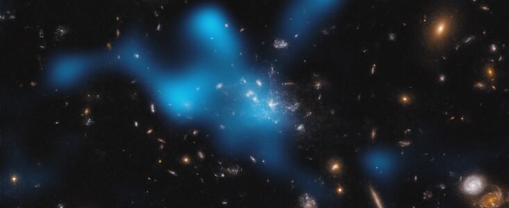 This image shows several galaxies distributed over a black background. The galaxies have colours that range from blue, orange-red, yellow and white. In the centre of the image is a larger concentration of galaxies. Overlaid on the galaxies in the middle is a blue and semi-transparent region. It has a clumpy form with indistinct contours. There are smaller, dark blue regions around it.