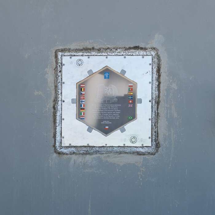 A grey concrete wall with a metal square box fitted into it — the time capsule. Etched into its surface is a hexagon with flags and wording too small to make out.