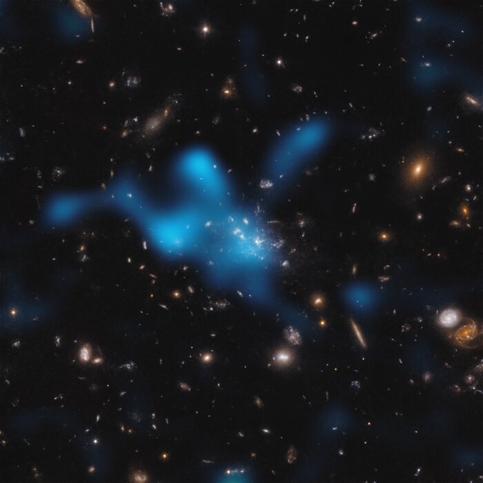 This image shows several galaxies distributed over a black background. The galaxies have colours that range from blue, orange-red, yellow and white. In the centre of the image is a larger concentration of galaxies. Overlaid on the galaxies in the middle is a blue and semi-transparent region. It has a clumpy form with indistinct contours. There are smaller, dark blue regions around it.