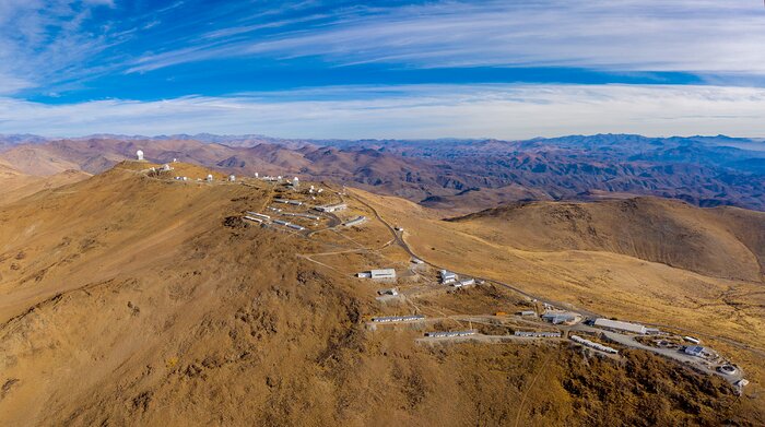 Many white domes are scattered across the road that follows the mountain ridge that makes up La Silla observatory. The mountainous desert that stretches in all directions have yellow-brown sand that contrasts to the blue sky.