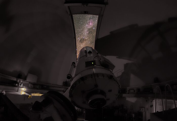 This picture is taken within the dome of the telescope, so most of this picture shows the telescope and the dome's interior. As it is nighttime, the dome is dark and the hatch is open, such that the night sky can be seen through the narrow hatch opening, out of which the telescope is pointing. The pink clouds of the Carina nebula, dark clouds, and bright stars are visible through the aperture.