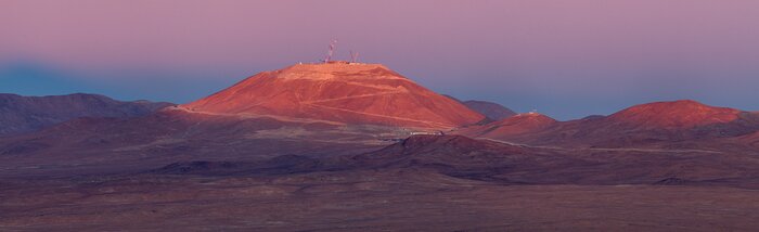 A landscape image of the desolate Atacama desert with the mountain, Cerro Armazones, in the centre at quite some distance from the photographer. The red desert is mostly in shadow, except for the top of Armazones, which is still illuminated by the setting sun. The service road and cranes are clearly visible and the only markers of human life. The sky is a beautiful pink that fades into dark blue towards the horizon.