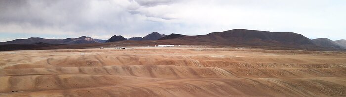 The image shows the vast, empty plains of the Chajnantor plateau in the Atacama Desert, Chile. The surroundings feel empty, other than a very small huddle of white structures at the centre of the image — these are the antennas of ALMA. Behind the dry, brown plains, mountain tops crop up on the horizon. Above the sky is grey and moody.
