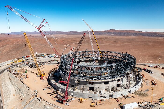 The image shows a construction site, littered with cranes, building materials and vehicles. The focal point of the image is undoubtedly the shell of what will one day be the ELT, circular in shape with a concrete base, topped off by the incomplete steel frame of the dome. It is truly colossal, dwarfing nearby cars by comparison. Behind the building site is the brown plains of the Atacama desert and in the distance mountains erupt from the ground, including Cerro Paranal, where the VLT resides. The sky above is a vibrant, electric blue.