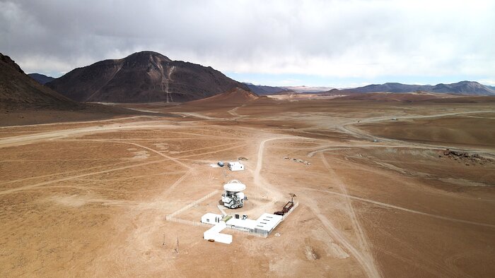 The image shows an open plain of a desert, with some white structures located centrally in the foreground. The white structures comprise a large dish, pointed skyward, and some surrounding ancillary buildings. The bright white of the technologically advanced man-made structures are in stark contrast to the brown plains of the Atacama desert that stretch off into the distance. On the left and in the distance on the right some mountains jut out from the flat plateau, while up above dark grey clouds stir in the sky.