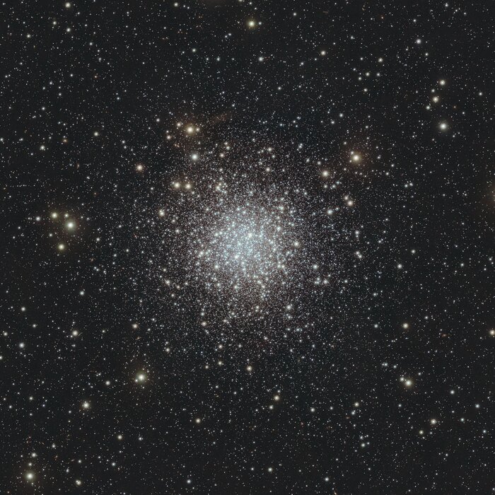 A dark background is dotted with countless white and yellow stars in this image, almost like snow falling on a winter’s night. At the centre, the density of these dots increases, forming a circular bright white region where the background is almost completely obscured. This dense region is the globular cluster.